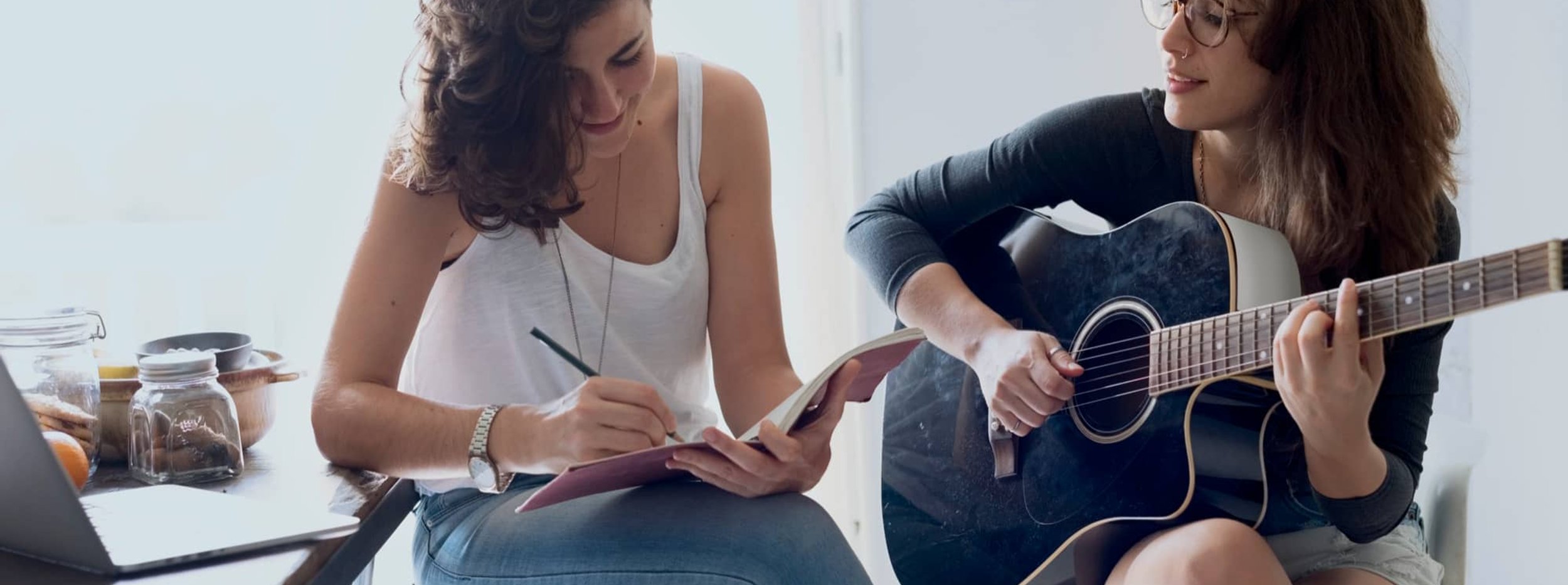 Woman playing guitar while another writes in notebook nearby
